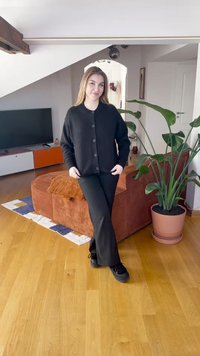 Svart strikket cardigan med knapper, kombinert med svarte bukser og svarte sko. Scenen har tregulv og en grønn plante i en potte.