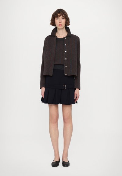 Chaqueta corta a rayas gris y marrón con cuello redondo, combinada con un top gris ajustado y una falda negra plisada con un cinturón de acento dorado.