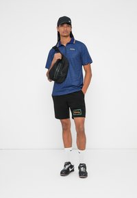 Polo a quadri blu con maniche corte, pantaloni corti neri con ricami floreali verdi e arancioni, borsa a tracolla nera e sneakers.