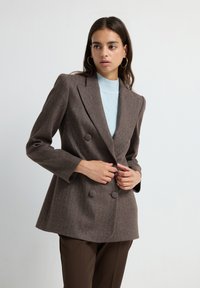 Blazer doppiopetto in tessuto a zigzag marrone con rever affilati, maniche lunghe e vestibilità rilassata, abbinato a un top azzurro in maglia.