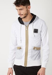 Chaqueta blanca con cremallera y capucha negra, que presenta dos bolsillos grises con cremallera y un detalle de cremallera dorada. Tejido texturizado con un parche de logo.