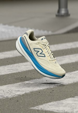 Beež ja sinine New Balance jooksujalats õhus asfaldist ülekäiguraja kohal, rõhutades kergesti kantavat disaini ja pehmendust.