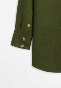 Olive green linen shirt with a rounded hem and buttoned cuffs. Features four buttons on the sleeve with a textured fabric finish.