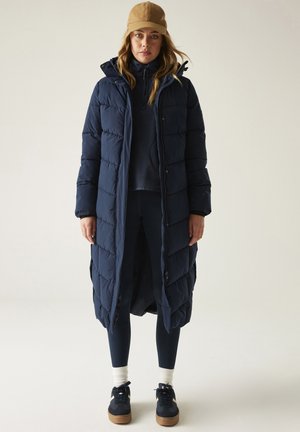 Donna in piedi che indossa un lungo piumino blu navy, leggings blu navy, sneakers blu navy con calzini bianchi, top blu navy e un cappellino da baseball beige.