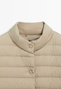 Giacca imbottita beige con collo alla coreana, caratterizzata da cuciture orizzontali, due bottoni a pressione e finitura liscia opaca.