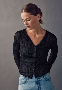 Schwarzes, texturiertes Hemd mit Knopfleiste, feinen wellenförmigen Linien, langen Ärmeln und V-Ausschnitt. Kombiniert mit hellblauen Jeans, die ein geschichtetes Design zeigen.