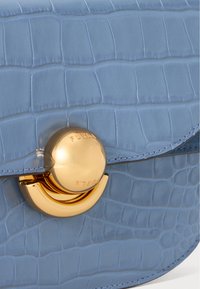 Light blue crocodile-embossed leather handbag with a round gold clasp featuring engraved branding, accent stitching, and a smooth finish.