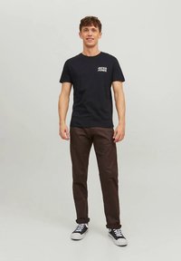 Juoda medvilninė marškinėliai su balta „JACK & JONES“ logotipu ant krūtinės, derinami su rudomis kelnėmis ir juodais sportbačiais su baltais padais.