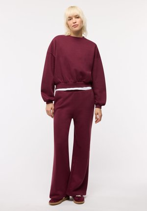 Giovane con capelli biondi che indossa una felpa bordeaux ampia e pantaloni a gamba larga abbinati, in piedi davanti a uno sfondo bianco.