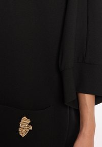 Schwarzes Kleidungsstück aus strukturiertem Stoff mit lockeren Ärmeln. Verfügt über eine dekorative goldene Hardware-Akzent auf der Tasche.