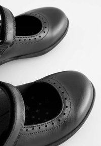 Pantofi Mary Jane din piele neagră cu detalii perforate și vârf rotund. Au o curea Velcro și o interior cu model în formă de stea.