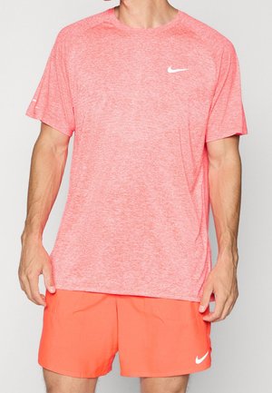 Man wearing coral pink Nike short-sleeve athletic shirt and bright orange Nike shorts standing against a plain background.