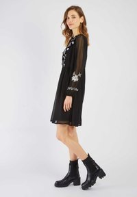 Zwarte doorzichtige jurk met bloemdecoratie langs de mouwen, gecombineerd met stevige zwarte enkellaarsjes met ribbeldetails en een dikke zool.