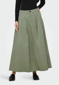 Une longue jupe plissée en tissu vert olive avec une fermeture à boutons à la taille et une forme trapèze, associée à des mocassins noirs.