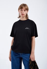 Camiseta de algodón negro de manga corta, con bordado blanco de la firma en el lado izquierdo del pecho. Combinada con pantalones de mezclilla azul.