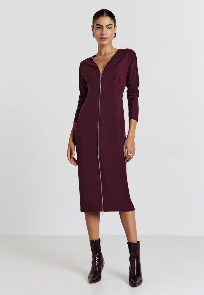 Frau in einem burgunderroten Midikleid mit langen Ärmeln und einem Reißverschluss vorne, kombiniert mit passenden Knöchelstiefeln, steht vor einem schlichten hellen Hintergrund.