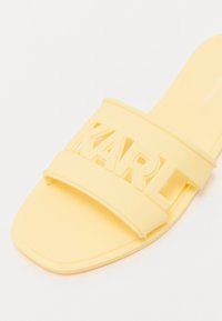Gula gummisandaler i sandaldesign med två breda remmar, med ett urklippt varumärke "KARL" i fet stil över framdelen. Platt sula.
