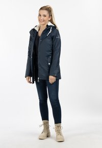 Giacca impermeabile blu navy con cappuccio foderato in pelliccia, chiusura con zip e automatici, due tasche frontali e cordini bianchi per la regolazione.