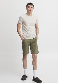 Bež majica kratkih rukava, maslinasto zelene šortse, bijele čarape i crne cipele na slip-on. Opusten kroj, pamuk s glatkom teksturom.