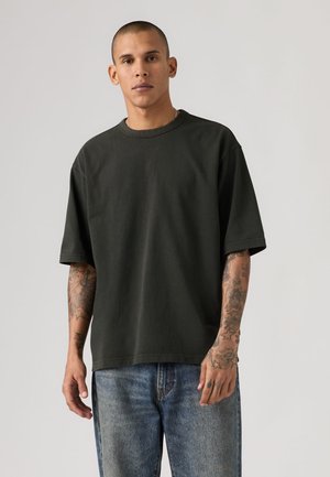 Joven con cabeza rapada que lleva una camiseta negra holgada y vaqueros azules, mostrando tatuajes detallados en ambos antebrazos y manos.