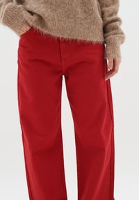 Pantalones de algodón rojos con un corte relajado, que presentan un diseño de cinco bolsillos y una textura sutil. Combinados con un suéter beige y esponjoso.