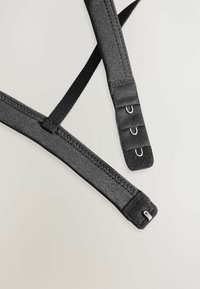 Zwarte elastische banden kruisen elkaar, met metalen haken en verstellers. De textuur is glad met een matte afwerking en subtiele stiksels.