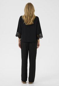Schwarze Bluse mit Spitzenakzenten an den Glockenärmel, kombiniert mit schwarzen Hosen. Glattes Material, schlichtes Design, von hinten getragen.