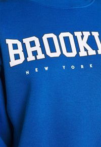Felpa blu con colletto a coste, con la scritta bianca in bold "BROOKLYN" e "NEW YORK" più piccola sotto, tessuto di una texture liscia.
