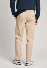 Chinos beige con design a gamba dritta, dotati di tasche posteriori con chiusura a bottone. Abbinati a una maglietta a maniche lunghe blu navy e sneaker sportive.