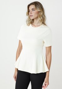 Top peplum bianco realizzato in tessuto morbido, con maniche corte, scollo tondo e orlo svasato, abbinato a pantaloni neri.