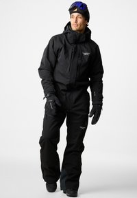 Zwarte waterdichte sneeuwoverall met een rits aan de voorkant, meerdere zakken en versterkte knieën. Inclusief handschoenen en een skielhelm met goggles.