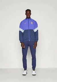 Mann som står, iført en blå og lilla sportsjakke med glidelås og emblem, marineblå bukser og hvite joggesko mot en enkel lys bakgrunn.