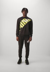 Felpa nera con grafica gialla diagonale e logo bianco, abbinata a pantaloni jogger neri con logo bianco. Scarpe nere con accenti verde.