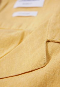 Primo piano di un tessuto giallo con cuciture visibili e due etichette bianche sfocate sullo sfondo.