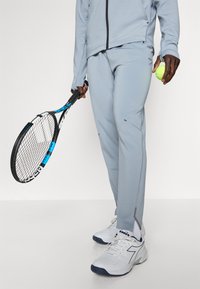 Pantalons de sport bleu clair avec des accents et coutures grises, accompagnés d'une raquette de tennis noire et d'une balle de tennis jaune, portés avec des baskets blanches.