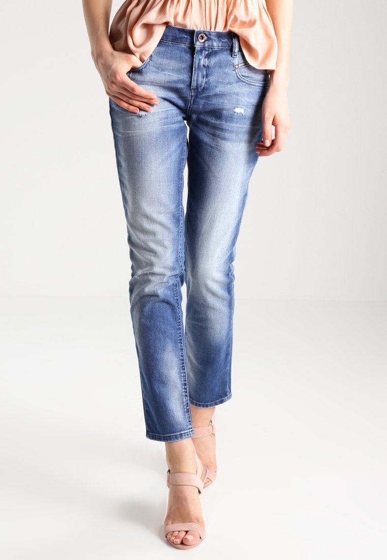 Vrouw gekleed in vervaagde blauwe jeans met lichte beschadigingen en een perzikblouse, poseert met één hand in de zak en roze hoge hakken.