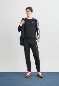 Melns sporta tērps ar baltām svītrām uz piedurknēm un biksēm, ar nelielu logotipu. Sarkanās sporta apavi papildina apģērbu.