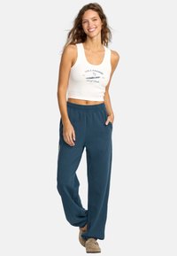 Vit crop top med blå grafisk text, tillsammans med löst sittande, teal sweatpants med elastiskt midjeband och fickor. Avslappnade skor synliga.