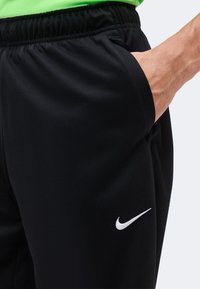 Personne portant un short noir Nike avec une taille élastique, main dans la poche, et logo blanc Nike swoosh visible sur la jambe.