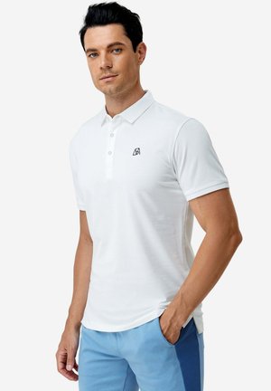 Mann trägt weißes Polo mit kurzen Ärmeln und kleinem schwarzem Logo auf der Brust sowie hellblaue Hose mit dunkelblauen Seitenstreifen, Hand in der Tasche.