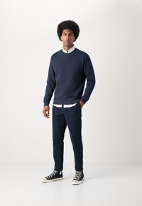 Navy sweater with round neckline, textured fabric, and small logo; paired with a white shirt, dark trousers, and black high-top sneakers.