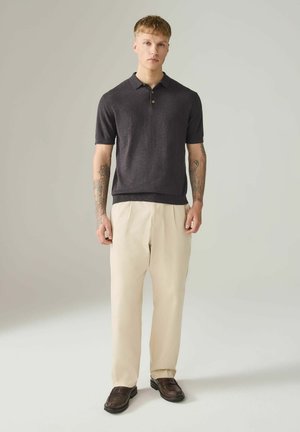 Junger Mann steht, trägt ein dunkelgraues kurzärmeliges Polo, hellbeige Hose, braune Loafers, mit Tätowierungen auf beiden Unterarmen vor einem schlichten Hintergrund.