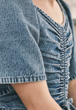 Haut en denim avec manches courtes bouffantes. Présente une texture froncée sur le corsage et un détail noué à la taille. Couleur bleu clair.
