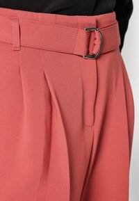 Pantalon corail taille haute avec plis et ceinture en tissu assorti attachée par une boucle en D argentée, associé à un haut noir.