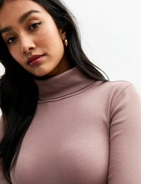 Top à manches longues et col roulé côtelé de couleur mauve, présentant un design ajusté et un col haut. Le modèle porte des boucles d'oreilles en or en forme de créoles.