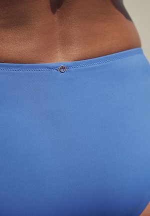 Primer plano de la parte baja de la espalda y la cintura de una persona que lleva un bañador azul con una pequeña hebilla metálica redonda en el centro.