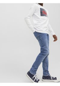 Camicia bianca a maniche lunghe con stampa grafica, jeans blu slim-fit e sneakers blu con suola bianca. Tessuto morbido e design casual.