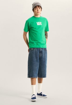 Zelené bavlněné tričko s bílým logem Vans, doplněné modrými džínovými kraťasy a modrými teniskami, zvýrazněné bílými kotníkovými ponožkami a šedou čepicí.