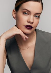 Złota biżuteria, głęboki burgundowy błyszczyk do ust i szary strój w serek. Tło jest neutralne, co podkreśla rysy twarzy i akcesoria.