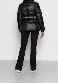 Osoba v černé prošívané bundě s logem Calvin Klein Jeans na zadní části pasu, v černých kalhotách a bílých teniskách, stojí čelem k boku.